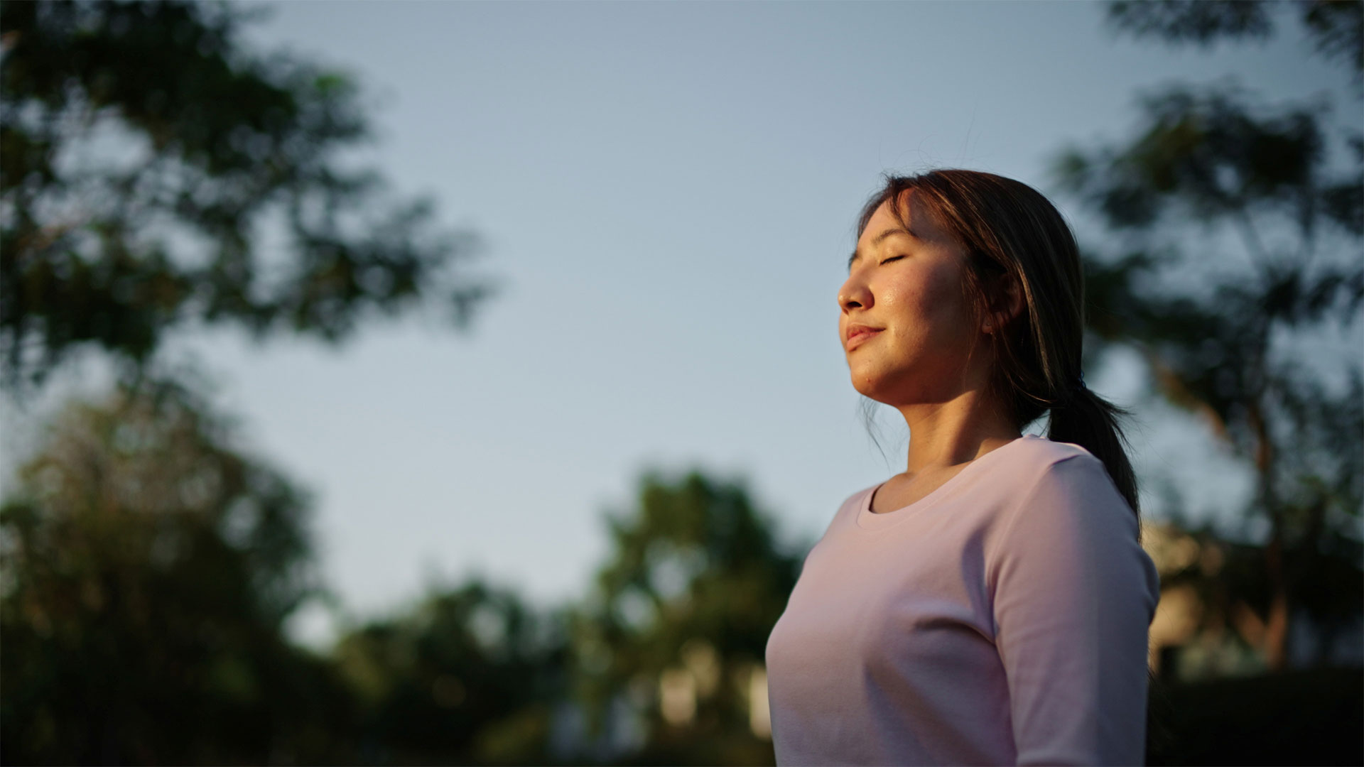 Person wearing a light pink long-sleeve top standing outdoors, with trees and greenery in the background under a clear sky, captured in soft natural light.