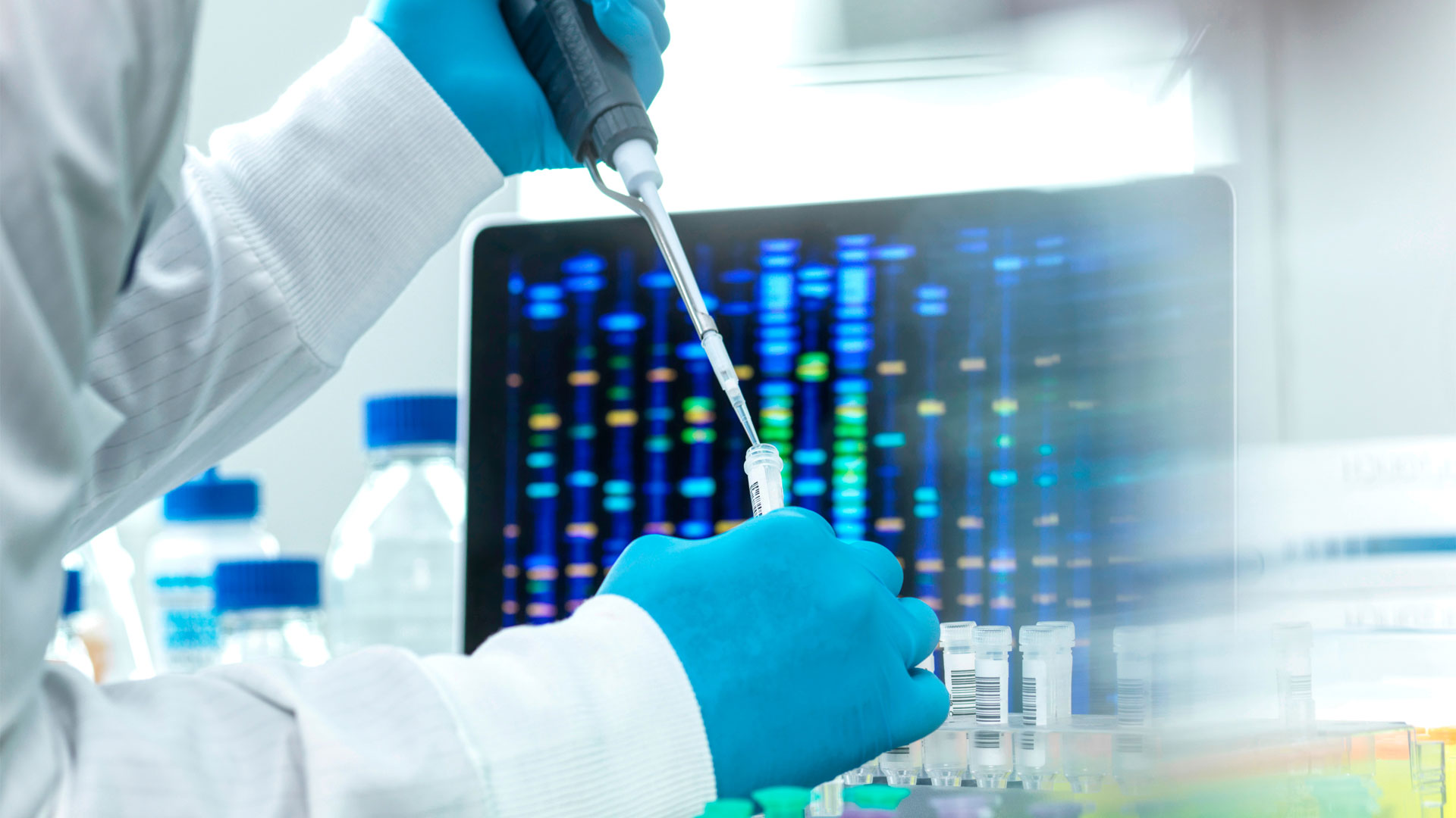 Scientist wearing blue gloves using a pipette to transfer liquid into a small tube in a laboratory, with a computer screen in the background displaying a colorful DNA gel electrophoresis pattern.