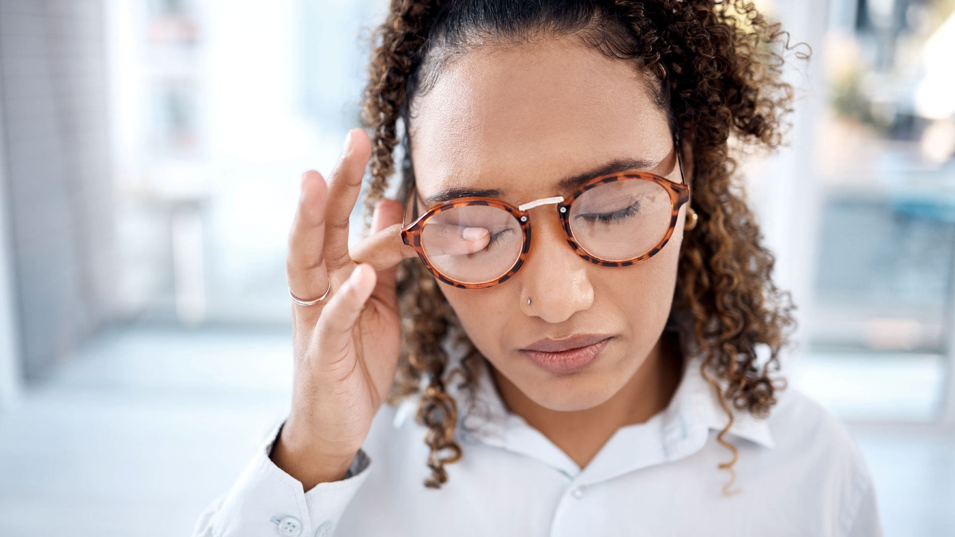 Person wearing a light-colored shirt indoors, rubbing eyes with one hand indicating tiredness, curly hair pulled back, and bright natural light in the background.